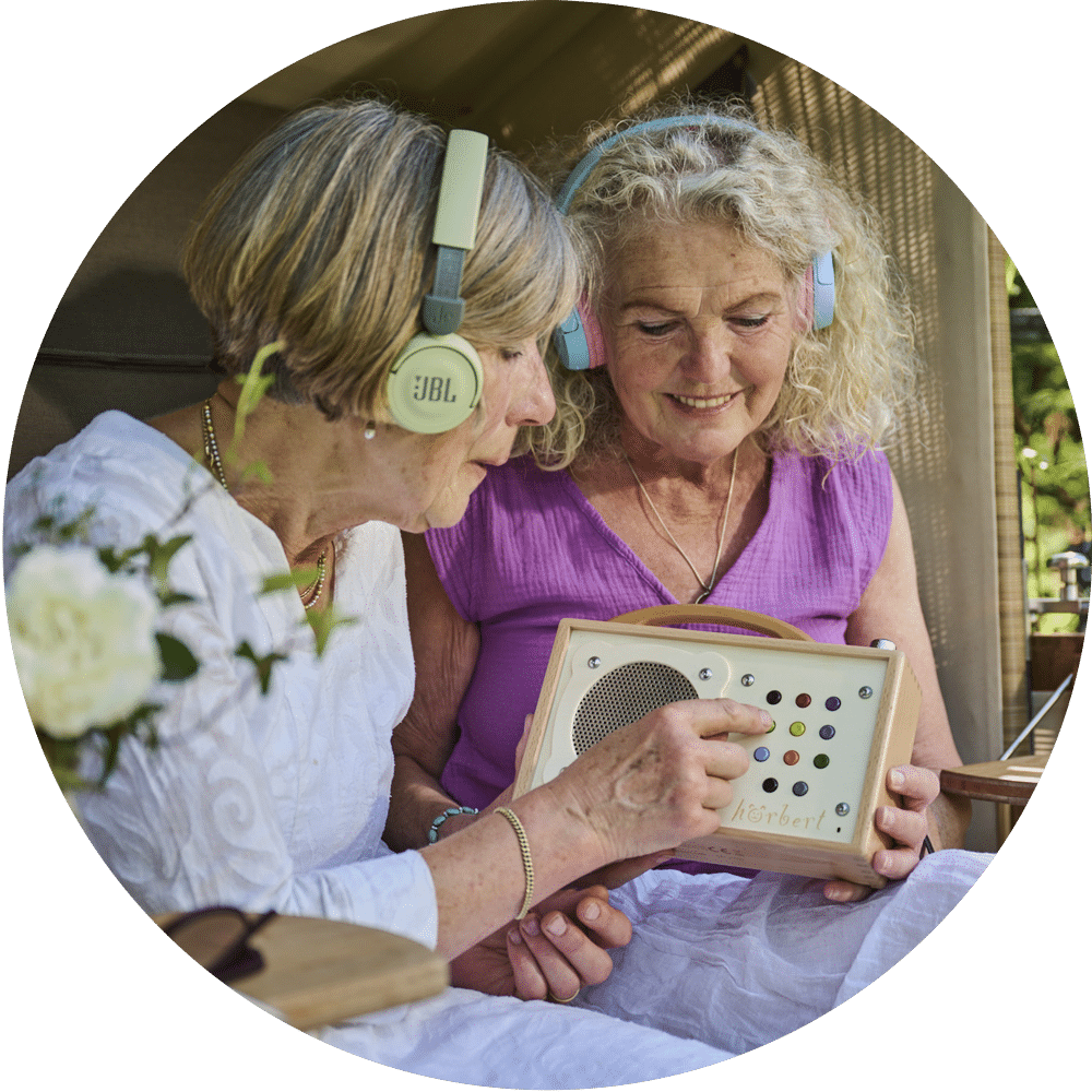 Zwei ältere Frauen, die Kopfhörer tragen, hören Musik über einen Hörbert-Lautsprecher.